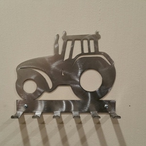 May include: Metal wall-mounted key holder shaped like a tractor with six hooks. The tractor is silver and has a brushed metal finish.