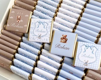 May include: A box of chocolates with gold foil wrappers. Some chocolates have light blue and white patterned wrappers, while others are brown with a teddy bear design. The name "Rohan" is printed on some of the chocolates.