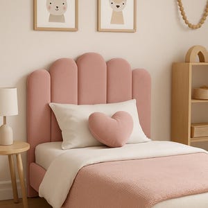 Puede incluir: Un marco de cama tapizado en color rosa con un cabecero festoneado, que incluye una almohada rectangular blanca y un cojín decorativo rosa en forma de corazón. La cama está vestida con una sábana blanca y una manta rosa con textura.