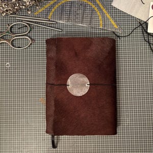 Könnte beinhalten: Ein handgefertigtes Notizbuch mit dunkelbraunem, strukturiertem Einband und einem silbernen, kreisförmigen Verschluss. Ein schwarzes Gummiband sichert das Notizbuch. Das Notizbuch hat ein schwarzes Leseband. Bastelwerkzeuge und -materialien sind im Hintergrund.