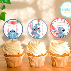 Puede incluir: Tres cupcakes con glaseado blanco y adornos de Stitch. Los adornos muestran el nombre "Olivia", la palabra "Aloha" y la frase "Thank for coming!" Los cupcakes están sobre una superficie de madera.