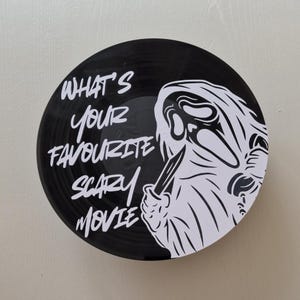 May include: A circular black vinyl record wall art piece featuring a white graphic illustration of a hooded figure holding a knife. The text "WHAT'S YOUR FAVOURITE SCARY MOVIE" is printed in white lettering on the left side of the record surface.