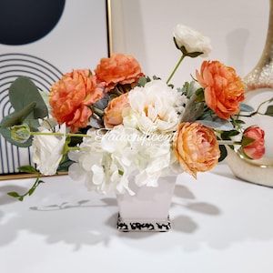 May include: A floral arrangement in a white square vase with a black and white patterned base. The arrangement features orange and white peonies, white roses, and green foliage. A decorative vase and artwork are in the background.
