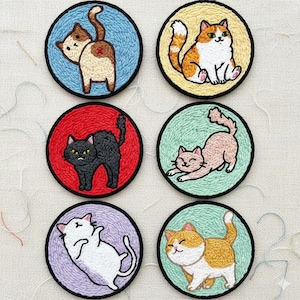May include: Six embroidered cat patches in a circular design. Each patch features a different cat illustration, with varying colours and poses, set against backgrounds of blue, yellow, red, green, and purple. The patches are outlined in black.