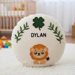 May include: A round, off-white pillow with embroidered details. Features the name "DYLAN" in black, a green four-leaf clover, green leafy branches, and a cartoon lion. The lion is brown, tan, and pink.