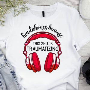 May include: White t-shirt with a red and white graphic of headphones. The text on the shirt reads "headphones because THIS SHIT IS TRAUMATIZING". The shirt is paired with blue jeans and white sneakers.