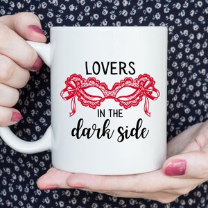 May include: White ceramic mug featuring the text "LOVERS in the dark side" in black script. A red lace mask design is centered above the text. The mug is held by a person with pink nail polish.