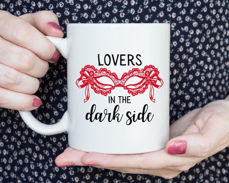 May include: White ceramic mug featuring the text "LOVERS in the dark side" in black script. A red lace mask design is centered above the text. The mug is held by a person with pink nail polish.