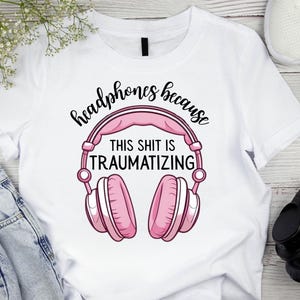 May include: White t-shirt featuring a pink and white graphic of headphones with the text "headphones because THIS SHIT IS TRAUMATIZING" in black script. The shirt is paired with blue jeans and white sneakers.