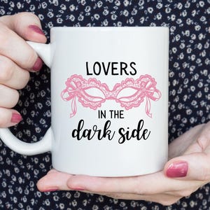 May include: White ceramic mug featuring the text "LOVERS in the dark side" with a pink lace mask design. The mug has a curved handle and is held by a person.