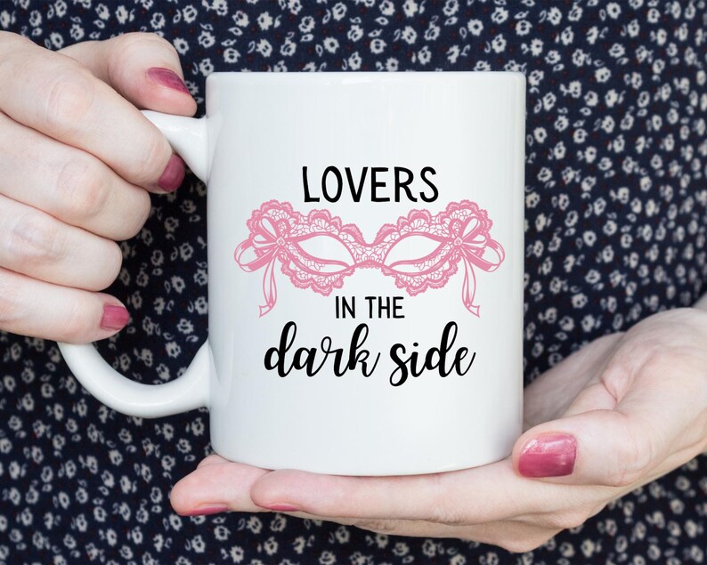 May include: White ceramic mug featuring the text "LOVERS in the dark side" with a pink lace mask design. The mug has a curved handle and is held by a person.
