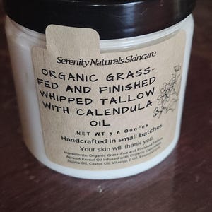 May include: A jar of Serenity Naturals Skincare whipped tallow with calendula oil. The label reads "Organic Grass-Fed and Finished" and "Net Wt. 3.6 Ounces." The jar has a black lid and a cream-colored label with text and a floral illustration.