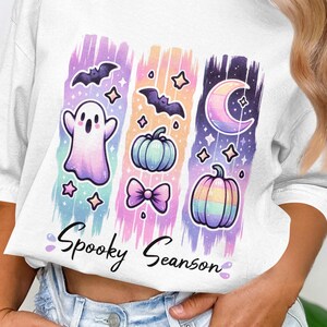 May include: White t-shirt with a colorful Halloween-themed graphic. The design features a ghost, bats, pumpkins, a bow, a crescent moon, and stars. The words "Spooky Seanson" are written in a script font.