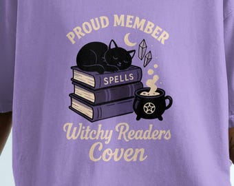 Könnte beinhalten: Ein lila T-Shirt mit einem Aufdruck, der eine schwarze Katze auf Büchern zeigt. Der Text lautet "Proud Member Witchy Readers Coven", mit Mond und Kristallen.
