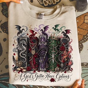 May include: A beige crewneck sweatshirt featuring a graphic print of five ornate, colorful books adorned with dragons, roses, chains, a crown, a dagger, and a key. Below the books, the text reads, A Girl's Gotta Have Options in a black script font.