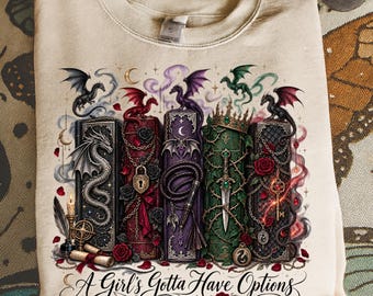 May include: A beige crewneck sweatshirt featuring a graphic print of five ornate, colorful books adorned with dragons, roses, chains, a crown, a dagger, and a key. Below the books, the text reads, A Girl's Gotta Have Options in a black script font.