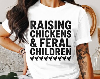 May include: White t-shirt with the text "RAISING CHICKENS & FERAL CHILDREN" in bold black letters. Below the text are black silhouettes of chickens. The shirt is a casual wear item.