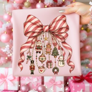 May include: A light pink fabric featuring a large, central red and white striped bow. Hanging from the bow are various Christmas-themed charms including a Santa Claus, a gingerbread person, a nutcracker, a snowman, a gift, and a tree.