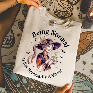 Pode incluir: Uma camisola bege de gola redonda com o gráfico de uma pessoa a usar um chapéu roxo de aba larga, segurando uma chávena de chá a fumegar. O texto "Being Normal Is Not Necessarily A Virtue" está impresso a preto à volta da ilustração.