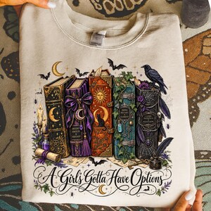 May include: A beige crewneck sweatshirt featuring a graphic print of five vintage books with celestial, floral, and occult motifs. The text "A Girl's Gotta Have Options" is printed in a decorative script font at the bottom of the illustration.