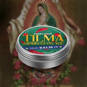 May include: A round, silver beard balm tin with a green label. The label reads "TILMA IMPRINTS ON YOU" in gold lettering, with "BEARD BALM" above. The brand name "Catholic BALM CO" is also visible. The background features a blurred religious image.
