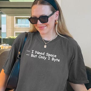 Peut inclure: T-shirt gris foncé avec le texte "- I Need Space - But Only 1 Byte" en blanc. Le t-shirt est de coupe ample et a des manches courtes. L'image comprend également un sac à main noir et des lunettes de soleil.