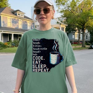 Peut inclure: T-shirt vert clair avec le texte "CODE. EAT. SLEEP. REPEAT." et des éléments de code HTML. Un graphique d'une tasse à café est sur le côté droit du t-shirt. Le t-shirt est ample.