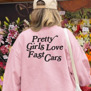 May include: Light pink sweatshirt with the text "Pretty Girls Love Fast Cars" in black. The sweatshirt is a soft pink color and has long sleeves. The image is of the back of the sweatshirt.