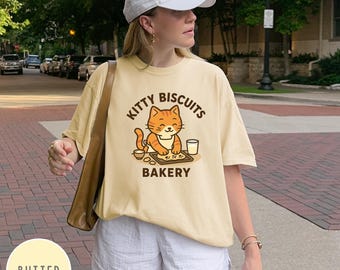 May include: Butter-colored t-shirt with a graphic of a smiling orange cat wearing an apron, with the text "Kitty Biscuits Bakery". The cat is depicted baking biscuits. The shirt is paired with white shorts and a white baseball cap.