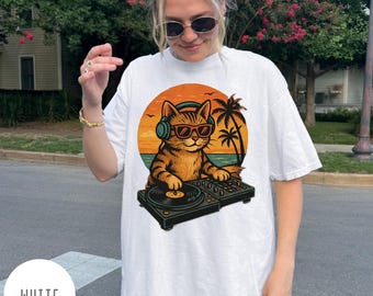 May include: White t-shirt featuring a cartoon cat DJ wearing sunglasses and headphones, set against a sunset beach scene with palm trees. The cat is operating a turntable. The word "WHITE" is printed at the bottom.