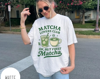 May include: White t-shirt with the text "Matcha Lovers Club" in green, along with illustrations of matcha drinks. The shirt also has the phrase "Ok, but first Matcha" in green. The shirt is worn by a person wearing sunglasses.