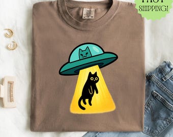 May include: Espresso-colored t-shirt featuring a cartoon design of a black cat being beamed up by a teal UFO. The UFO has a cat pilot. The shirt is on a white surface with gold earrings and a hair clip.