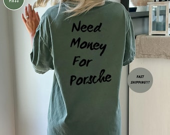 May include: A moss green t-shirt with the text "Need Money For Porsche" in black. The shirt is a relaxed fit and has short sleeves. The shirt is a solid color and has a casual style.