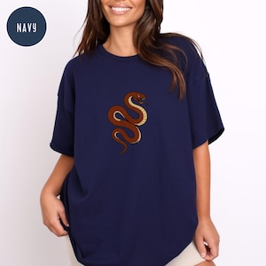 May include: Navy blue t-shirt featuring a detailed illustration of a brown snake with yellow accents. The snake is coiled and has a red head. The word "NAVY" is in a navy blue circle in the upper left corner.