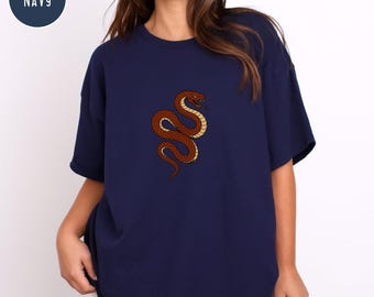 May include: Navy blue t-shirt featuring a detailed illustration of a brown snake with yellow accents. The snake is coiled and has a red head. The word "NAVY" is in a navy blue circle in the upper left corner.