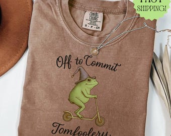 May include: Espresso-colored t-shirt with a whimsical graphic of a green frog wearing a wizard hat and riding a scooter. The text reads "Off to Commit Tomfoolery". A silver necklace is draped across the shirt.