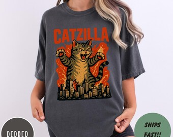 May include: Dark grey t-shirt featuring a cartoon cat graphic with the text "CATZILLA" in orange. The cat is depicted roaring and destroying a city, with flames in the background. The shirt is a comfortable, casual style.