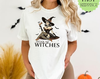 May include: White t-shirt featuring a graphic of a witch holding a cocktail, with the text "Drink up WITCHES". The witch is wearing a black hat and dress. Black bats are in the background. A green "FAST SHIPPING!" sticker is in the upper right corner.