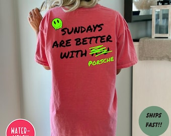 May include: A watermelon-colored t-shirt with the text "SUNDAYS ARE BETTER WITH PORSCHE" in black and neon green. The shirt features a neon green smiley face and a green scribble. The shirt is a casual style.