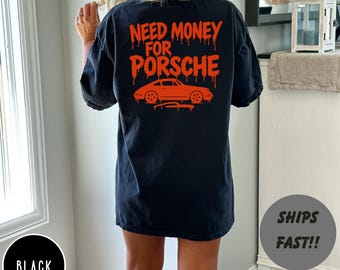May include: Black t-shirt with the text "NEED MONEY FOR PORSCHE" in orange, along with an orange car graphic. The text has a dripping effect. The shirt is a dark color.