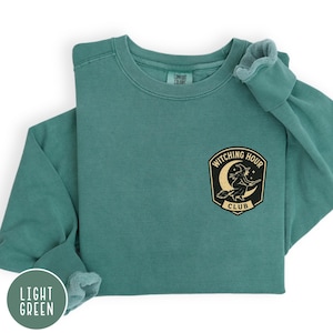 May include: A light green sweatshirt with the text "Witching Hour Club" on a patch. The patch features a witch on a broomstick in front of a crescent moon. The sweatshirt is a solid color and has a crew neck.
