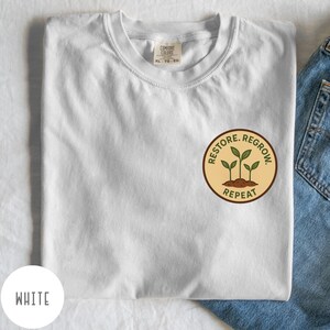 May include: White t-shirt with a crew neck and a circular graphic on the chest. The graphic features three green sprouts, the words "RESTORE. REGROW. REPEAT" in a brown circle. The shirt is folded, with a portion of blue denim jeans visible.