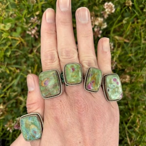 Könnte beinhalten: Nahaufnahme einer Hand mit sechs silbernen Ringen. Jeder Ring hat einen großen, quadratischen grünen und roten Edelstein. Die Ringe haben einen strukturierten Silberrand, vor grünem Gras.