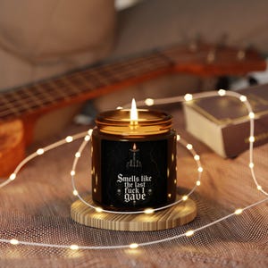 Pode incluir: Uma vela acesa num frasco de vidro âmbar com um rótulo preto apresentando um gráfico de uma vela e o texto "Smells like the last fuck I gave" em letras góticas brancas, acentuado por duas pequenas ilustrações de caveiras nas laterais.