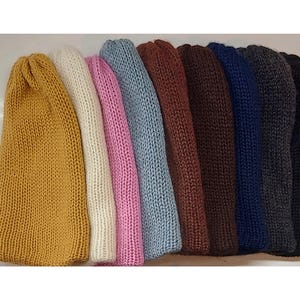 May include: A collection of nine knitted beanies arranged in a row, featuring a variety of solid colors including mustard yellow, cream, pink, light blue, brown, dark brown, navy blue, charcoal gray, and black in a ribbed texture.