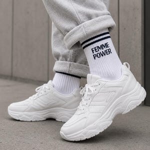 Puede incluir: Zapatillas blancas gruesas con un diseño texturizado. Las zapatillas se combinan con calcetines blancos con el texto "FEMME POWER" y pantalones de chándal grises. Los zapatos tienen una suela gruesa y cierre de cordones.