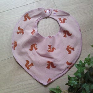 Könnte beinhalten: Hellrosa Baby-Lätzchen mit einem sich wiederholenden Muster aus orangefarbenen Eichhörnchen. Das Lätzchen hat eine abgerundete Form mit einem Druckknopfverschluss. Das Lätzchen ist aus weichem Stoff.
