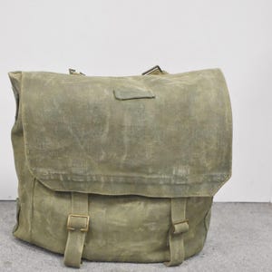 May include: An olive green canvas backpack with a flap closure and adjustable straps. The bag has metal buckles and a rectangular shape. The canvas material has a worn, textured appearance, suggesting age and use. The bag is designed for carrying items.