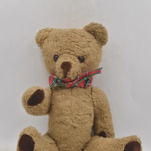 Puede incluir: Un oso de peluche marrón con ojos y nariz marrón oscuro, que lleva una pajarita a cuadros roja, verde y blanca. El oso tiene almohadillas de color marrón oscuro en sus patas y pies. El juguete es de felpa suave.