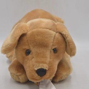 Puede incluir: Un peluche de perro golden retriever con ojos y nariz negros, y orejas caídas. El perro está tumbado con un calcetín blanco y rojo a rayas en la boca. El juguete está hecho de material suave y afelpado.